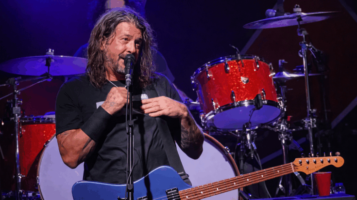 Dave Grohl stands onstage with his blue guitar, speaking into the microphone in front of a bright red drum kit.
