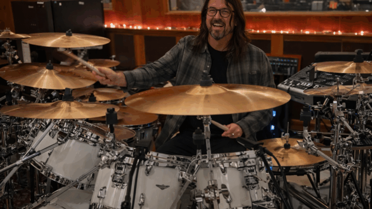 Dave Grohl smiles behind a large drum kit as he plays in a warm studio setting.