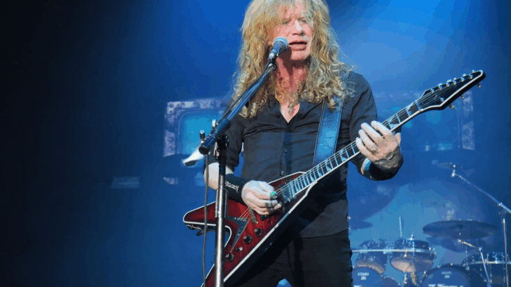 Dave Mustaine performs onstage under blue lights while playing a sharp-bodied electric guitar at the microphone.