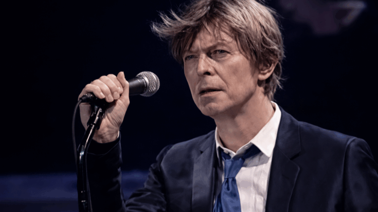 David Bowie stands at a microphone, gazing intensely as he prepares to sing under stage lighting.