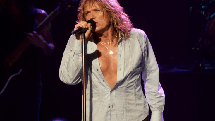 David Coverdale is singing into a microphone onstage, wearing an open shirt under dim concert lighting.