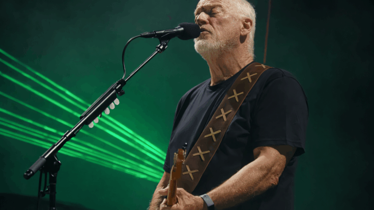 David Gilmour sings into a microphone while playing his guitar under green stage lights.