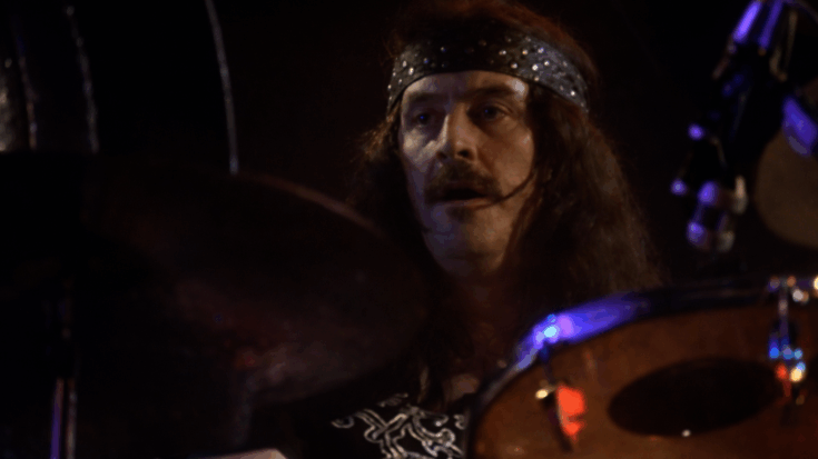 John Bonham looks intense behind his drum kit, framed by dark stage lighting and a studded headband.