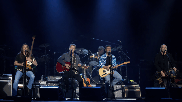 The Eagles perform live onstage under blue lights, with the band seated and playing guitars around the drummer.