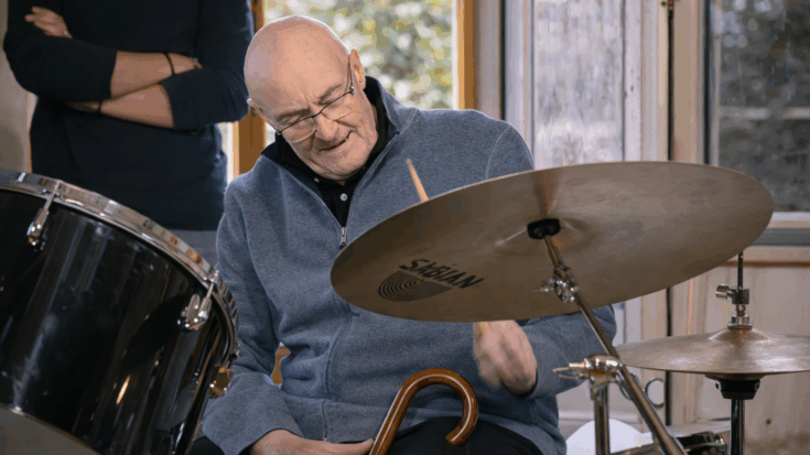 Phil Collins smiles as he plays the drums, holding a cane while seated at his kit in a relaxed indoor setting.