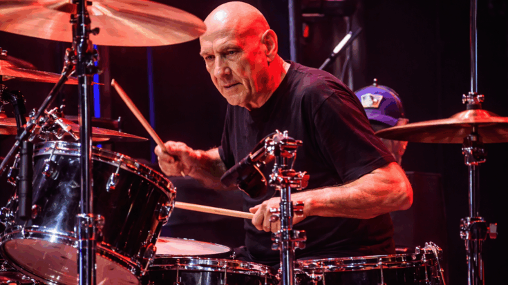 Bill Ward plays the drums with fierce focus under vivid red stage lights.
