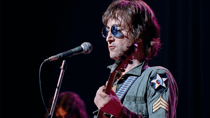 John Lennon is depicted performing onstage in his iconic olive drab military jacket and blue-tinted round glasses, holding a guitar while positioned in front of a microphone.