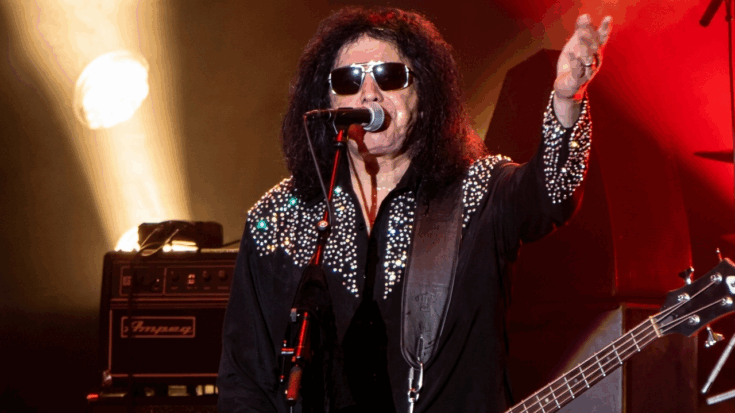 Gene Simmons performs on stage in a studded black shirt and sunglasses, raising one hand while playing his bass guitar during a live set.