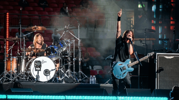 Foo Fighters performing on stage, with Dave Grohl singing and pointing upward while playing a guitar, and the late Taylor Hawkins playing the drums in the background.