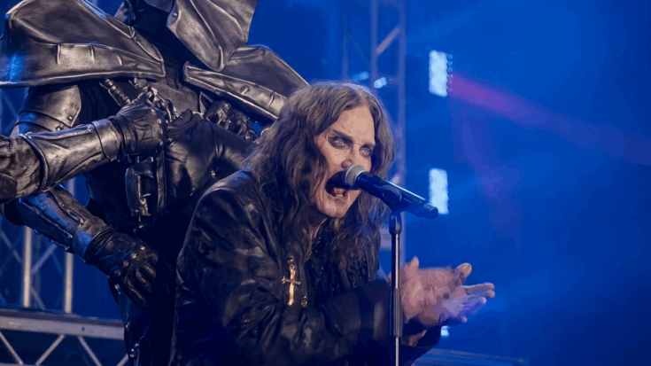 Ozzy Osbourne performing live, singing into a microphone with his hands raised, dressed in his characteristic black jacket with a large gold cross.