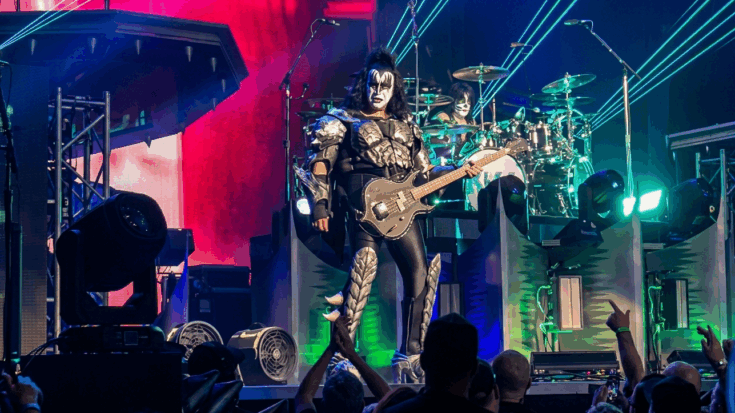 Gene Simmons stands center stage in full Demon regalia, playing his signature axe bass during a high-energy live performance.