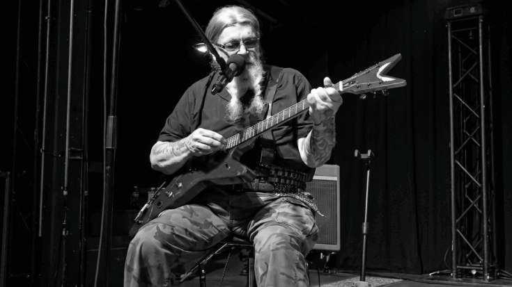 David Allan Coe performing on stage, playing a red electric guitar and singing into a microphone while seated.