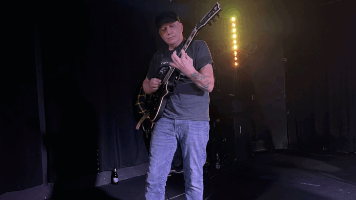 Chris Poland plays his electric guitar with intense focus on a dimly lit stage illuminated by a vertical string of lights.