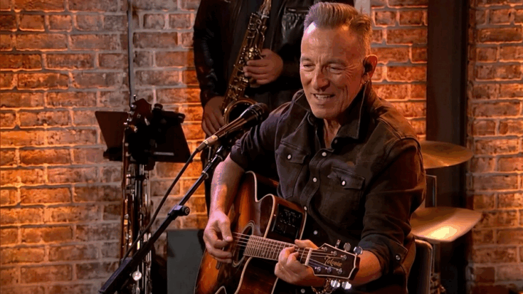Bruce Springsteen is captured in a warm, intimate moment, smiling while playing an acoustic Takamine guitar in front of a brick wall and a backing band.