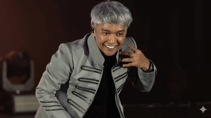 Arnel Pineda is seen smiling with silver-grey hair while wearing a grey military-style jacket featuring black horizontal stripes over a black shirt.