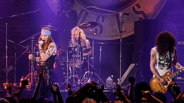 Guns N' Roses is captured in a high-energy live performance, featuring Axl Rose singing at the microphone and Slash playing his iconic Les Paul guitar under moody purple stage lights.