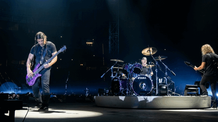 Metallica performs on a massive stage, with Robert Trujillo on bass and Kirk Hammett on guitar flanking Lars Ulrich's central drum kit.