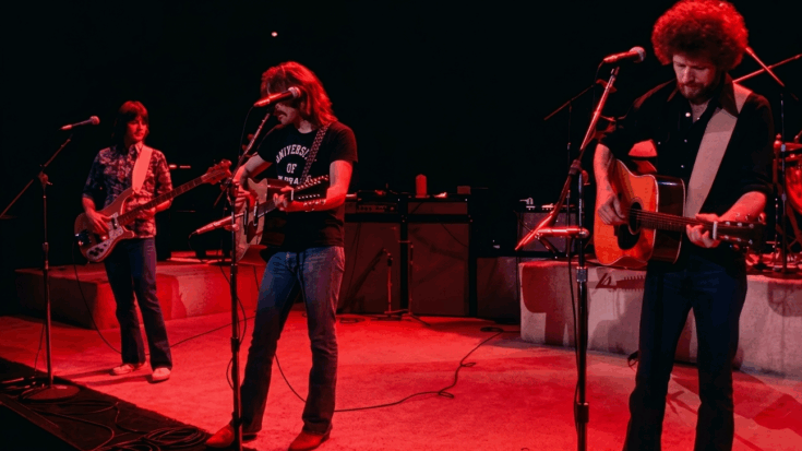 Eagles performing live on a dimly lit, red-washed stage, featuring two acoustic guitarists in the front and a bassist on the left.