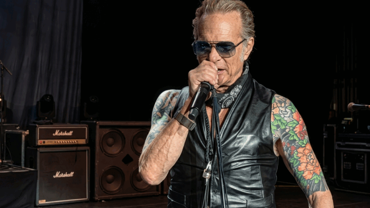 David Lee Roth is shown singing into a microphone during a performance, wearing a black leather vest, sunglasses, and a bandana that highlights his colorful floral tattoos.