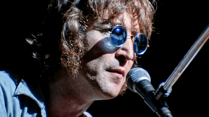 John Lennon sings into a microphone, wearing his signature round, blue-tinted sunglasses during a live performance.