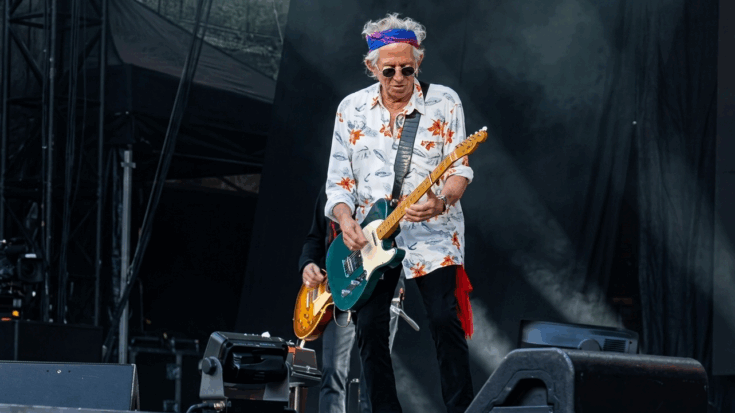 Keith Richards is captured performing onstage with a teal Fender Telecaster, wearing a floral shirt, dark sunglasses, and a colorful headband.