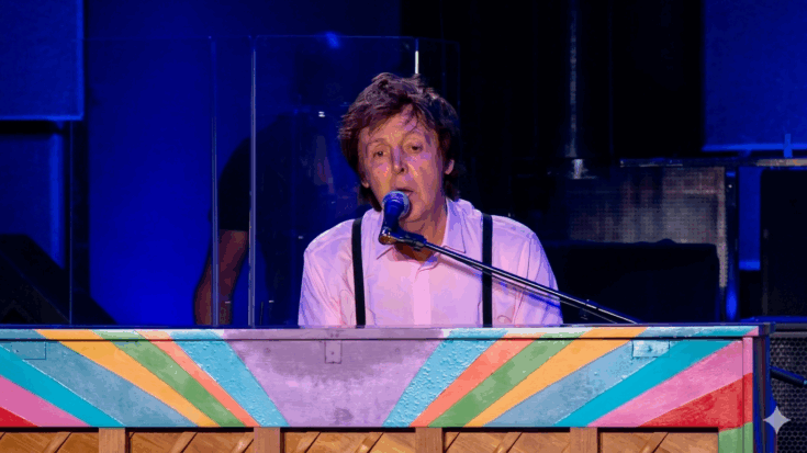 Paul McCartney is seen performing at a colorful, striped piano while wearing a light pink shirt and black suspenders.
