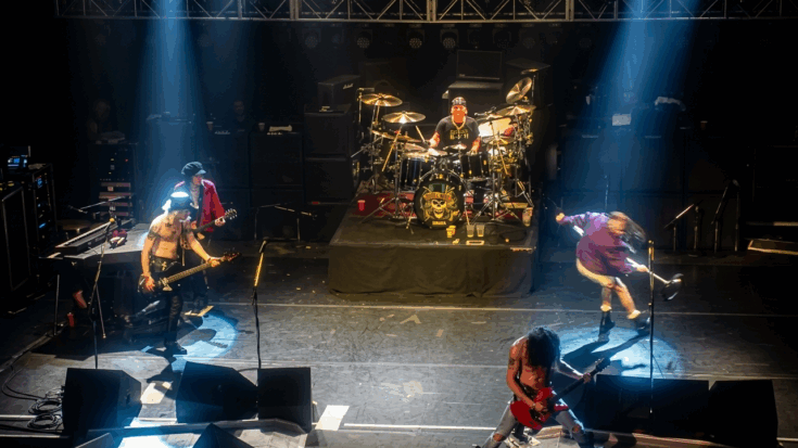 Guns N' Roses performs live on stage under spotlights, featuring Slash playing guitar in the foreground and a prominent band logo on the drum kit.