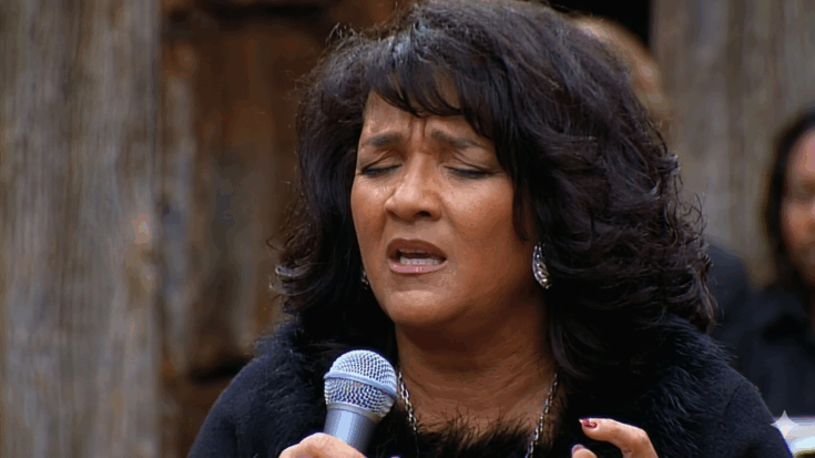 Nedra Talley Ross is captured singing with her eyes closed, wearing a black fur-trimmed top and silver jewelry while holding a microphone.