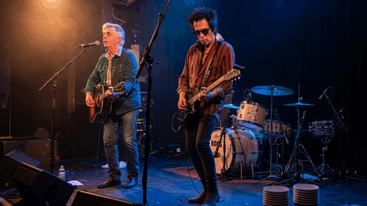 Earl Slick plays a black electric guitar alongside a singer on an intimate stage, wearing sunglasses and a striped shirt during a live performance.