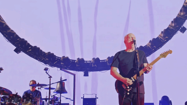 David Gilmour is shown performing center stage with his black Fender Stratocaster in front of the iconic circular light rig and a massive projection screen.