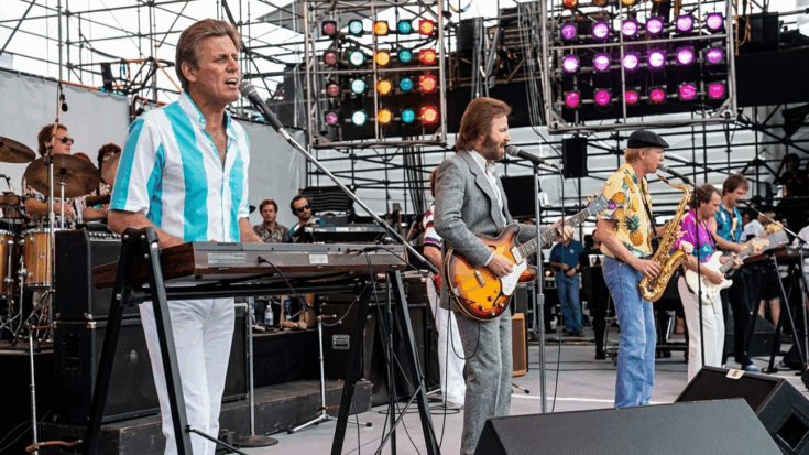 The Beach Boys perform on an outdoor stage with Mike Love at the keys and Brian Wilson on guitar, backed by a full band in vibrant, tropical attire.