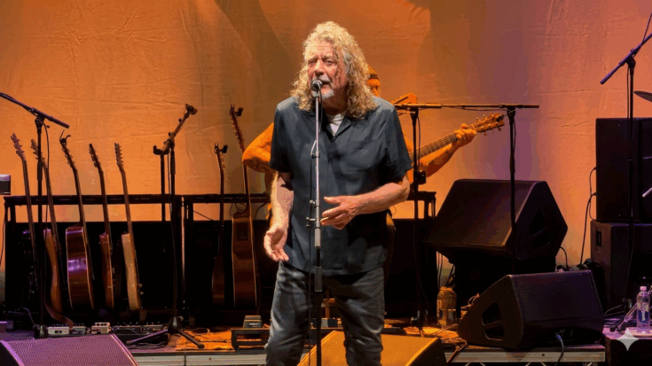 Robert Plant is performing on stage with long curly hair, wearing a dark short-sleeved button-down shirt while singing into a microphone.