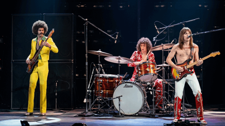 Grand Funk Railroad performs a high-energy set on stage, featuring a bassist in a bright yellow suit, a drummer in patterned red attire, and a shirtless guitarist leading the power trio.