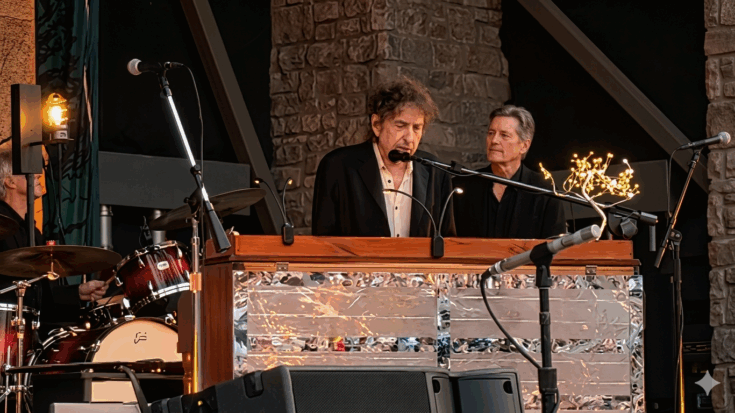 Bob Dylan is seen performing at a piano on stage alongside other musicians, wearing a dark suit jacket over a light-colored shirt.