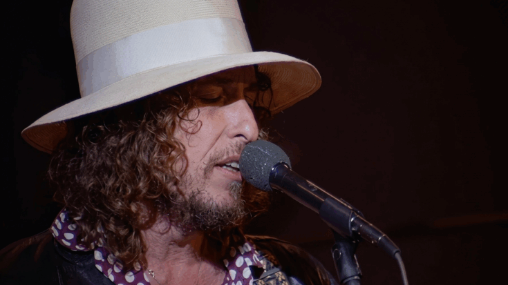 Bob Dylan is shown in a tight profile shot from the Rolling Thunder Revue era, wearing his signature wide-brimmed white hat and singing intently into a microphone.