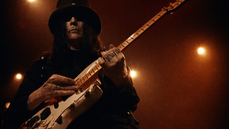A low-angle shot captures Mick Mars in his signature black hat, intensely playing a worn electric guitar under warm stage lights, giving him a dark and powerful presence.