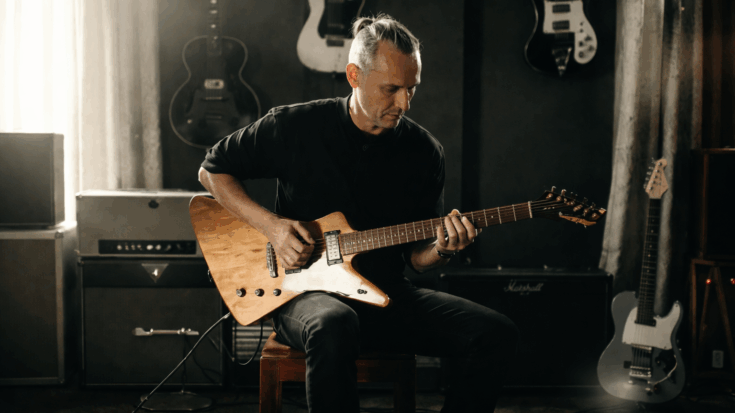 Tom Dumont sits in a studio, focused on playing guitar in a calm and intimate setting.