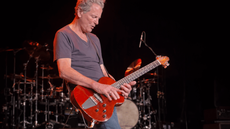 Lindsey Buckingham plays a bright red electric guitar onstage under warm red lighting.