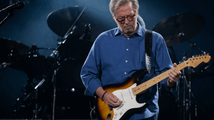 Eric Clapton stands onstage under soft blue lighting, calmly focused as he plays his sunburst electric guitar.