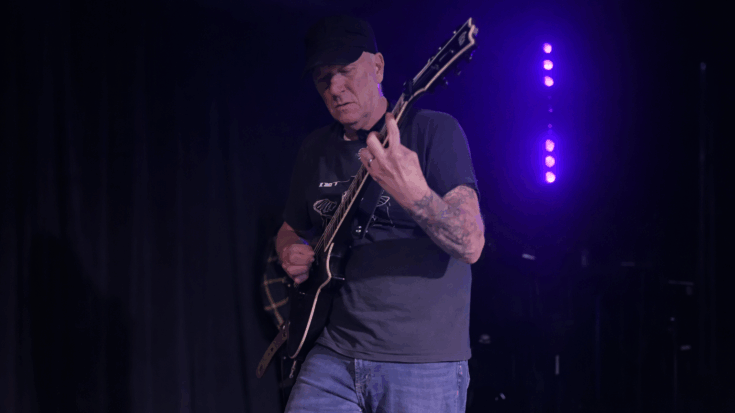 Chris Poland plays guitar onstage under purple lighting, focused on his instrument in a quiet, intimate performance moment.