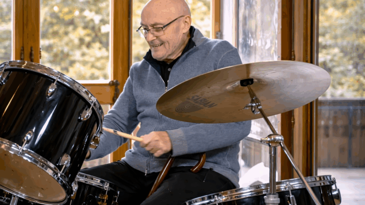Phil Collins smiles as he plays the drums, captured mid-performance in a relaxed, joyful moment.