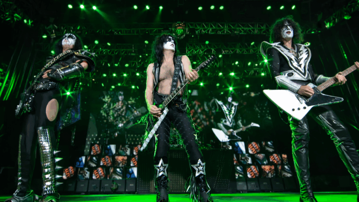 KISS performs on stage in full makeup and iconic costumes, playing guitars under bright green concert lights.