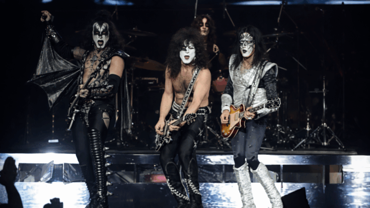 KISS is performing onstage in full makeup and costumes, playing guitars under bright concert lights.