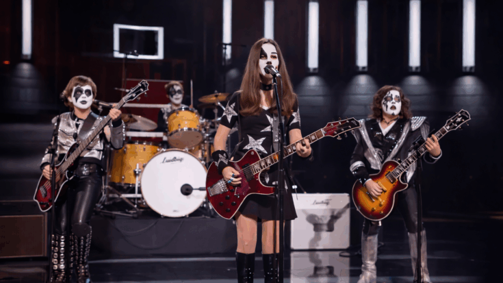 A young rock band performs onstage in KISS-style makeup and costumes under bright concert lights.