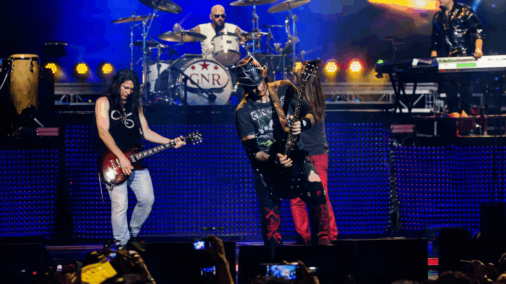 Guns N’ Roses are performing live on a brightly lit stage with each band member locked into the moment.