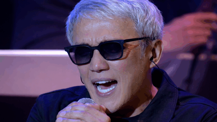 Arnel Pineda is shown in a close-up performance shot, singing into a microphone while wearing dark sunglasses.