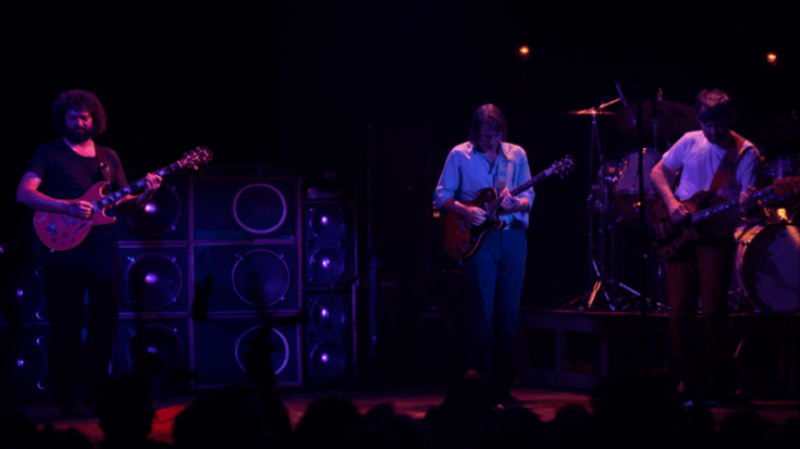 Grateful Dead are performing live on a dimly lit stage, with the band members playing guitars while the drummer keeps rhythm in the background.