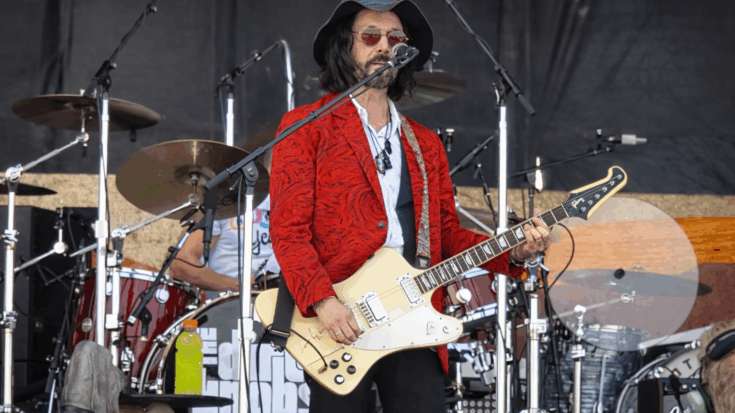 Mike Campbell performs on stage in a red jacket and hat, playing guitar and singing into a microphone.