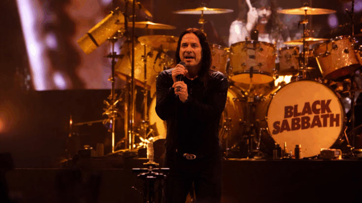 Black Sabbath is performing live onstage with the singer Ozzy Osbourne at the front and the drummer playing behind a kit marked with the band’s name.