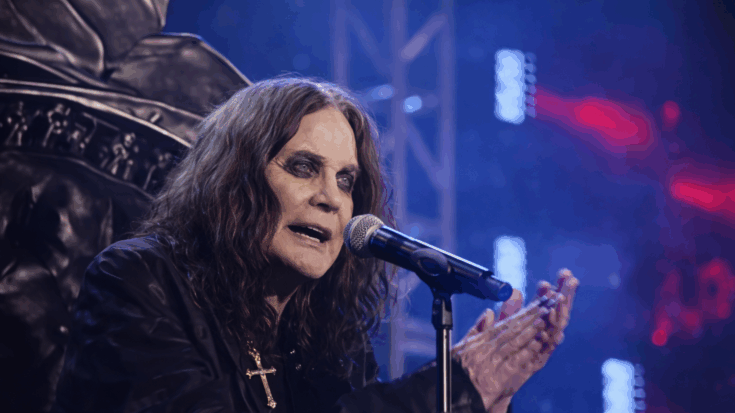 Ozzy Osbourne stands onstage at a microphone, gesturing with his hands under blue concert lighting.
